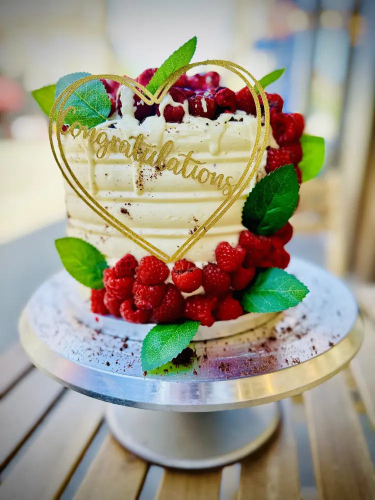 Tall cake with white buttercream, fresh raspberries, mint leaves and a gold Congratulations topper